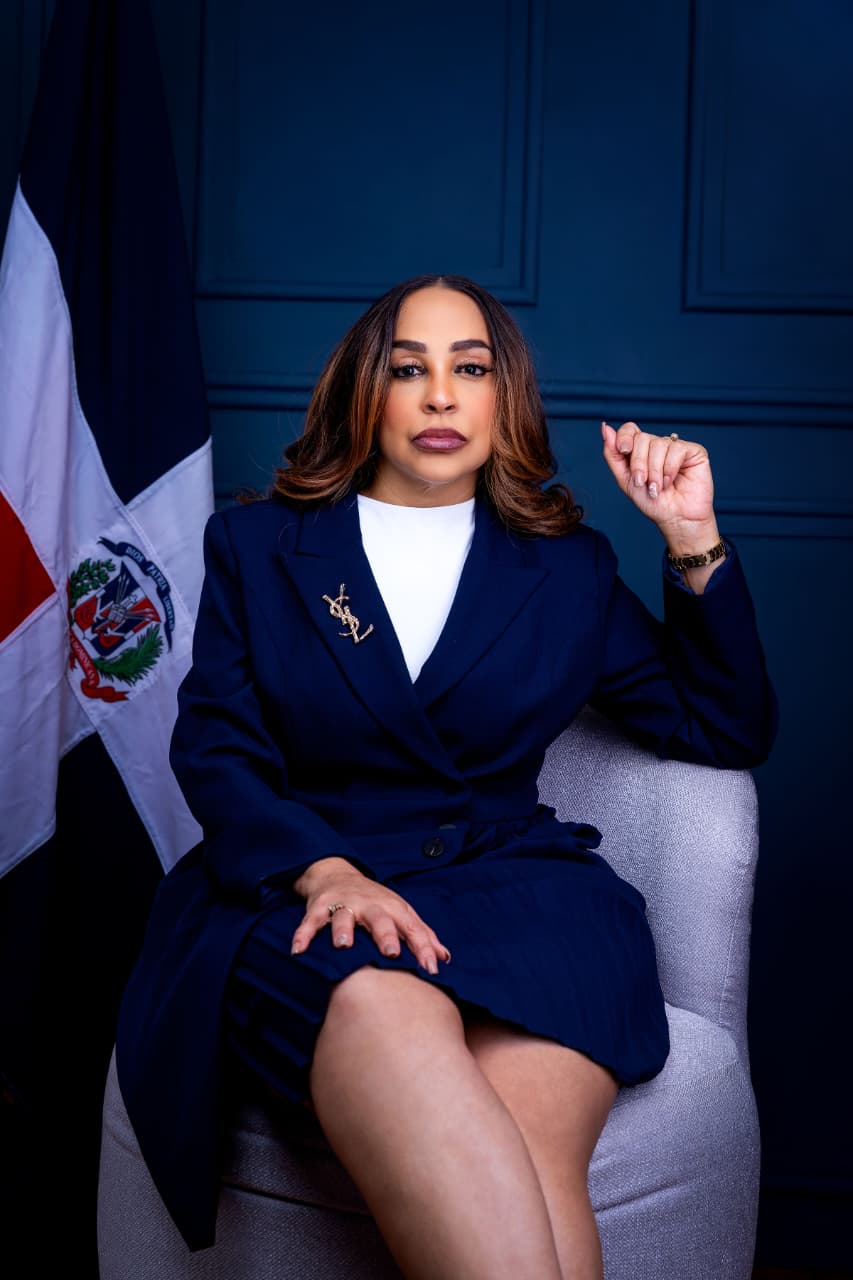 Katy Báez sentada, con chaqueta azul marino, junto a la bandera dominicana